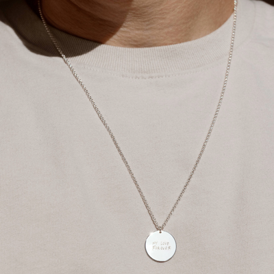 A lifestyle photo of a person's neck wearing a silver chain necklace with a round silver pendant engraved with handwritten script.
