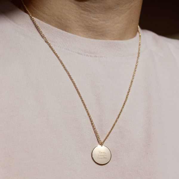 A lifestyle photo of a person's neck wearing a gold chain necklace with a round gold pendant engraved with handwritten script.