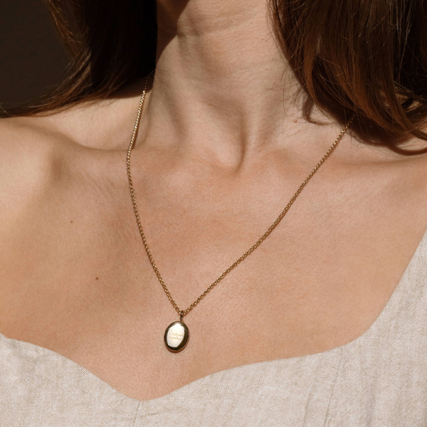 A lifestyle photo of a person's neck wearing a gold chain necklace with a gold oval locket pendant engraved with handwritten script.