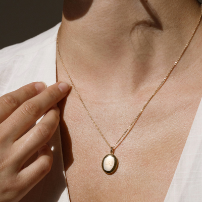 A lifestyle photo of a person's neck wearing a gold chain necklace with an oval gold locket pendant engraved with handwritten script.