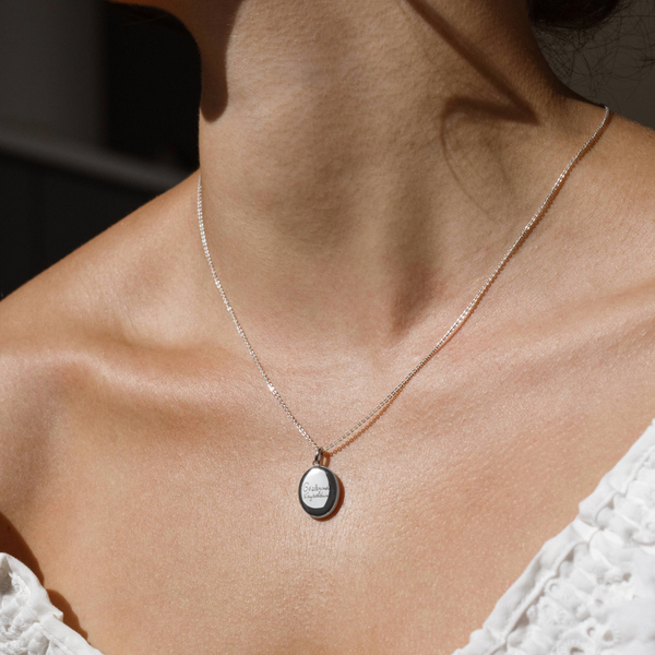 A lifestyle photo of a person's neck wearing a silver chain necklace with a silver oval locket pendant engraved with handwritten script.