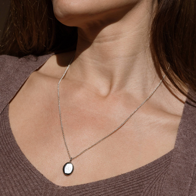 A lifestyle photo of a person's neck wearing a silver chain necklace with a silver oval locket pendant engraved with handwritten script.