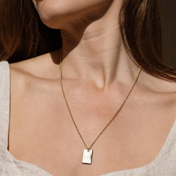 A close up lifestyle photo of a person wearing a dainty gold pendant necklace with a rectangular pendant, engraved with text, and a light colored top.