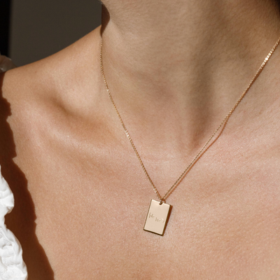A lifestyle photo of a person's neck wearing a gold chain necklace with a rectangular gold pendant engraved with handwritten script.