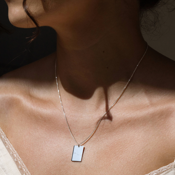 A lifestyle photo of a person's neck wearing a silver chain necklace with a rectangular silver pendant engraved with handwritten script.