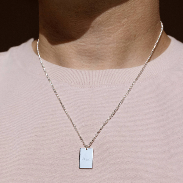 A lifestyle photo of a person's neck wearing a silver chain necklace with a rectangular silver pendant engraved with handwritten script.