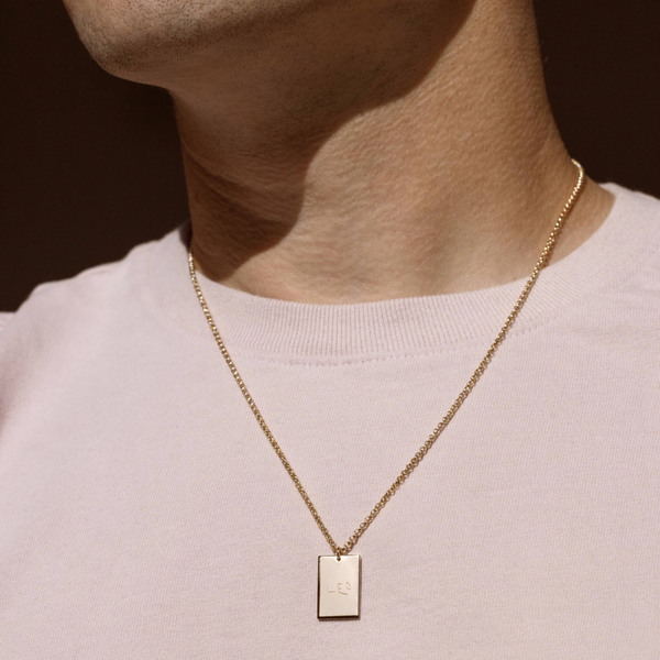 A lifestyle photo of a person's neck wearing a gold chain necklace with a rectangular gold pendant engraved with handwritten script.