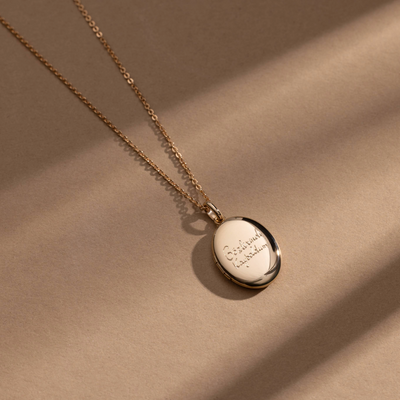 A gold chain necklace with an oval gold locket pendant engraved with handwritten script, shown against a solid light beige background.