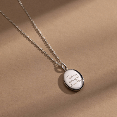 A silver chain necklace with a silver oval locket pendant engraved with handwritten script, shown against a solid light beige background.