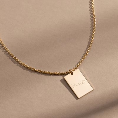 A gold chain necklace with a rectangular gold pendant engraved with handwritten script, shown against a solid light beige background.