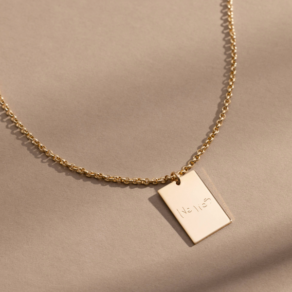 A gold chain necklace with a rectangular gold pendant engraved with handwritten script, shown against a solid light beige background.