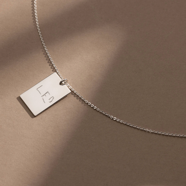 A silver chain necklace with a rectangular silver pendant engraved with handwritten text, shown against a solid light beige background.