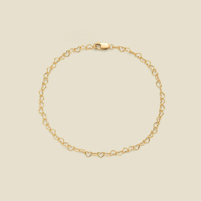 A delicate gold chain bracelet with a continuous open-heart link pattern and a lobster clasp, displayed on a solid light beige background.