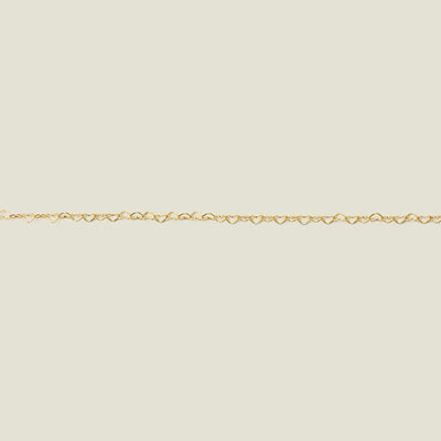 A gold heart link chain bracelet stretched horizontally across a solid light beige surface.