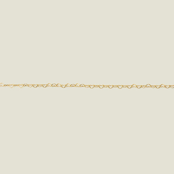 A gold heart link chain bracelet stretched horizontally across a solid light beige surface.