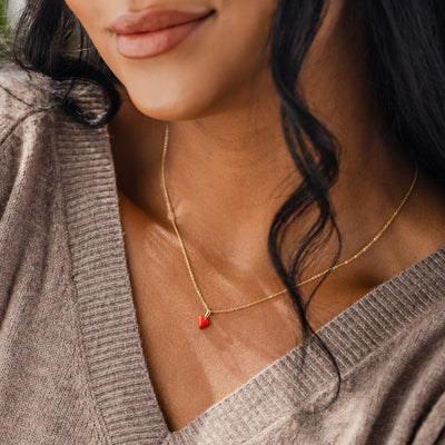 A close-up of a person wearing a grey v-neck knit top and a delicate gold chain necklace with a small red heart charm.