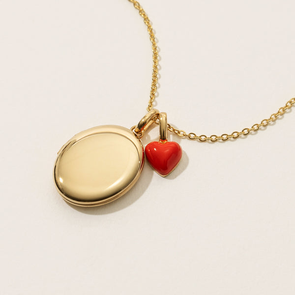 A gold circular link chain necklace with a gold oval locket charm and a small red heart charm, shown on a warm white background.