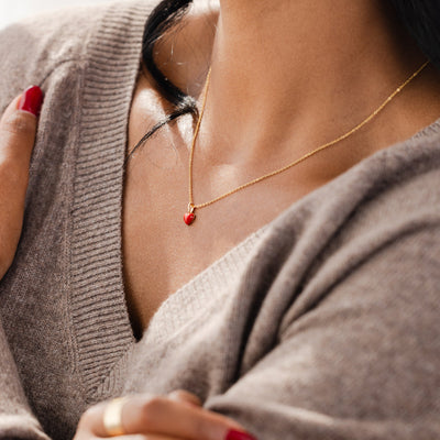 A close-up of a person wearing a grey v-neck knit sweater and a delicate gold chain necklace with a small red heart charm.