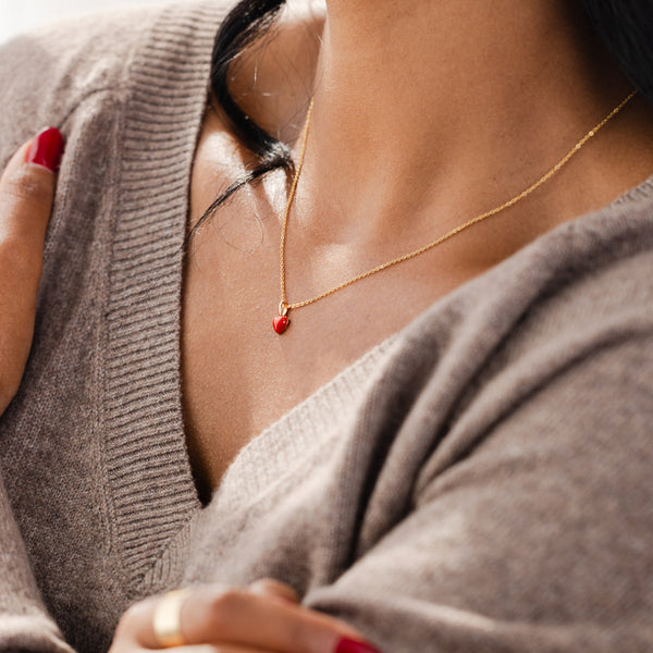 A close-up of a person wearing a grey v-neck knit sweater and a delicate gold chain necklace with a small red heart charm.