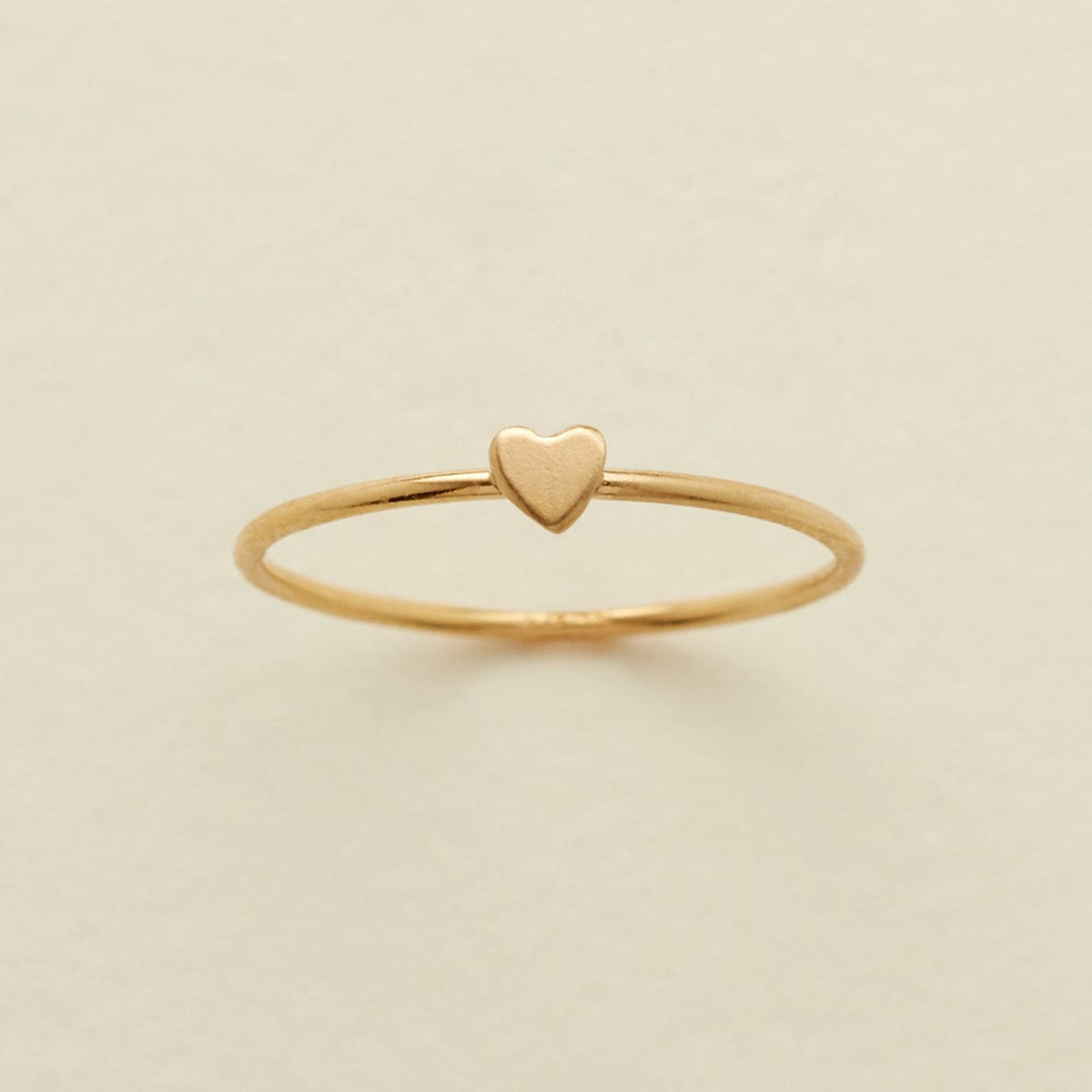 A delicate gold ring featuring a small, flat heart charm at the center, displayed on a soft, light beige background.