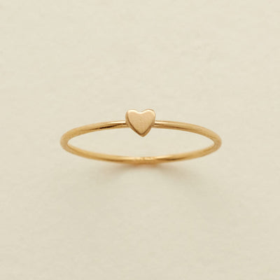A delicate gold ring featuring a small, flat heart charm at the center, displayed on a soft, light beige background.