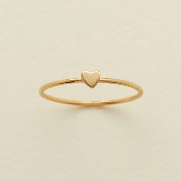 A delicate gold ring featuring a small, flat heart charm at the center, displayed on a soft, light beige background.