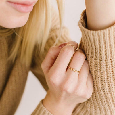 A person's hand wearing multiple thin wire gold rings, including one ring with a small flat gold heart in the center.
