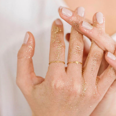 Two hands held together covered in gold glitter, wearing a thin gold wire ring and a thin gold wire ring with a flat heart in the center.