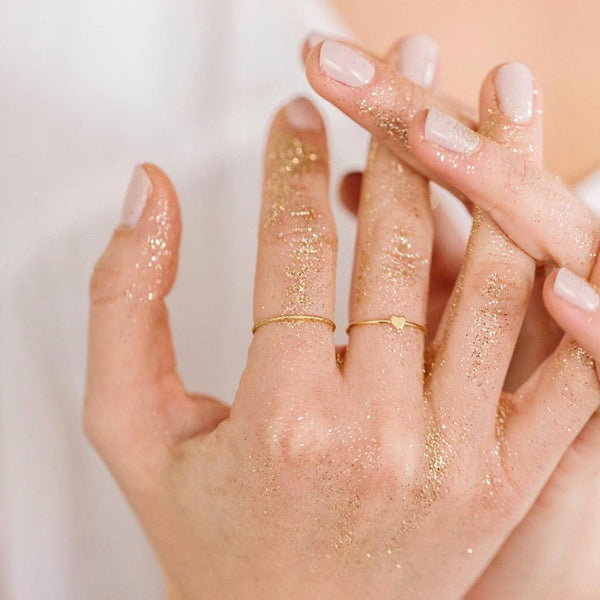 Two hands held together covered in gold glitter, wearing a thin gold wire ring and a thin gold wire ring with a flat heart in the center.