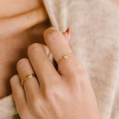 A hand resting on a chest with three stacked minimalistic gold wire rings on one finger and a thin gold wire ring with a flat heart in the center on another.