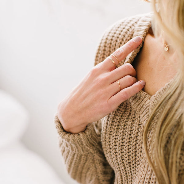 A hand resting on a chest with three stacked minimalistic gold wire rings on one finger and a thin gold wire ring with a flat heart in the center on another.