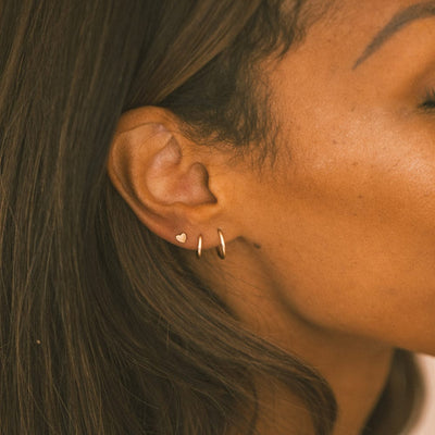 A close-up of a person's ear wearing two thin gold hoop earrings and a polished flat gold heart stud.
