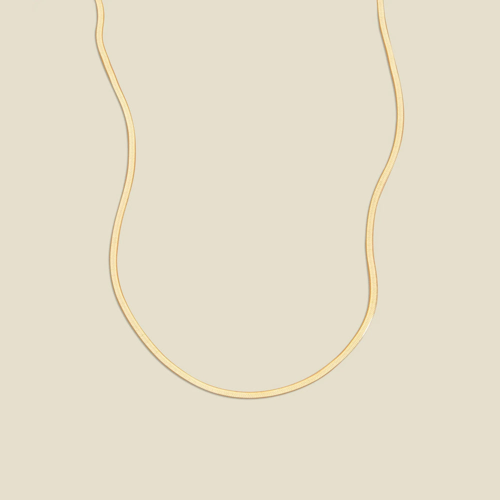A gold flat wide necklace, displayed on a solid light beige background.