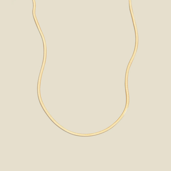 A gold flat wide necklace, displayed on a solid light beige background.
