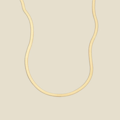 A gold flat wide necklace, displayed on a solid light beige background.