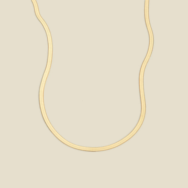 A gold flat wide necklace, displayed on a solid light beige background.