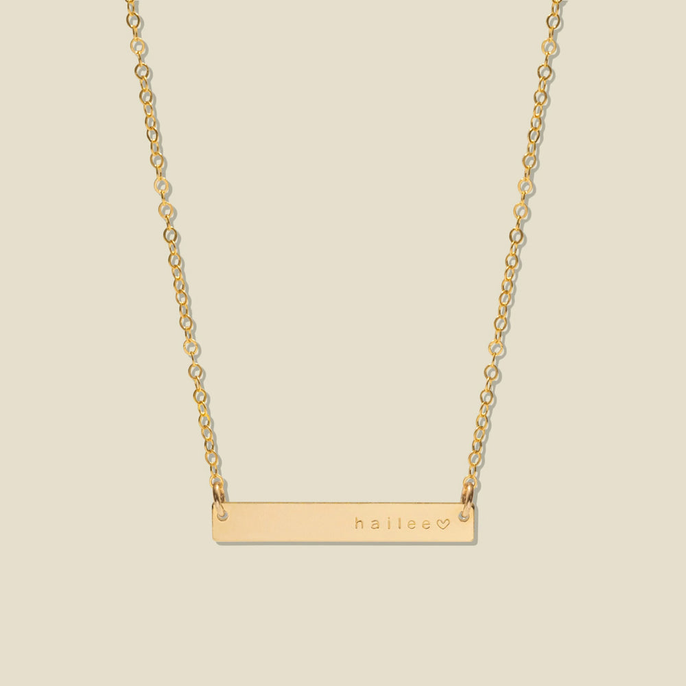 A gold bar necklace with a delicate chain, featuring the name 'haliee' and a small heart engraved on the right side of the bar, set against a solid light beige background.