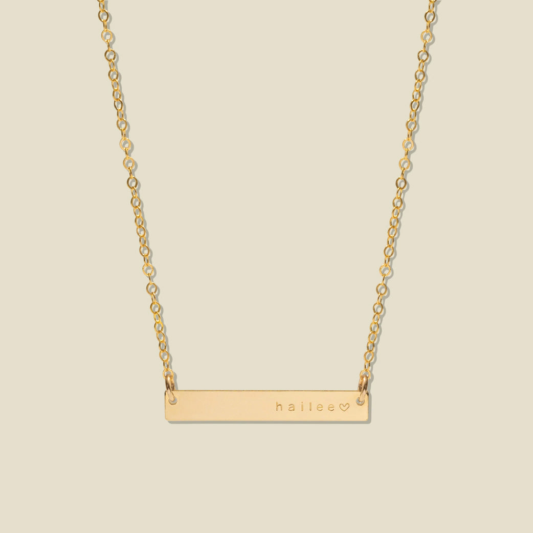 A gold bar necklace with a delicate chain, featuring the name 'haliee' and a small heart engraved on the right side of the bar, set against a solid light beige background.
