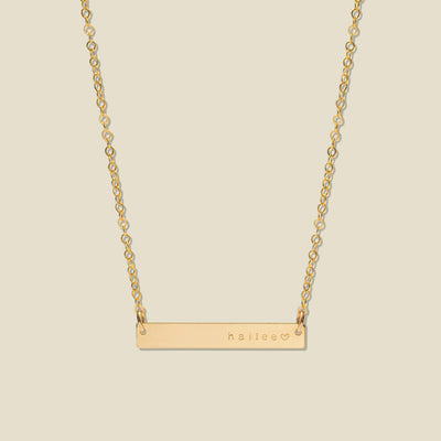 A gold bar necklace with a delicate chain, featuring the name 'haliee' and a small heart engraved on the right side of the bar, set against a solid light beige background.