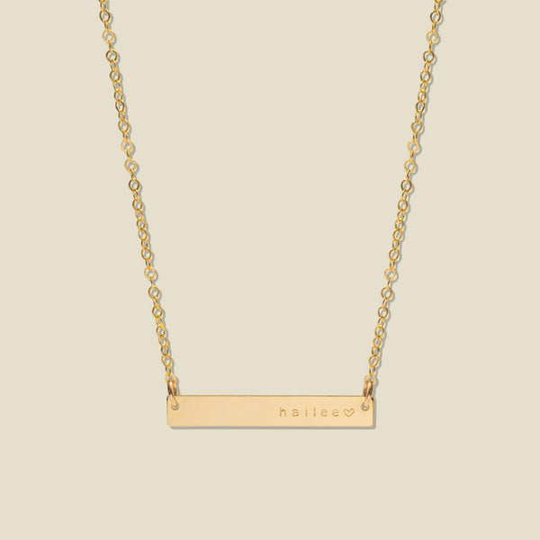 A gold bar necklace with a delicate chain, featuring the name 'haliee' and a small heart engraved on the right side of the bar, set against a solid light beige background.