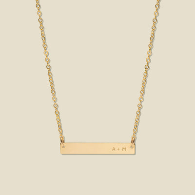 A gold bar necklace with a delicate chain, featuring 'A + M' engraved on the right side of the bar, set against a solid light beige background.