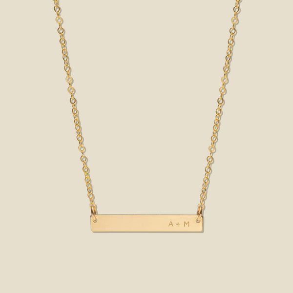 A gold bar necklace with a delicate chain, featuring 'A + M' engraved on the right side of the bar, set against a solid light beige background.