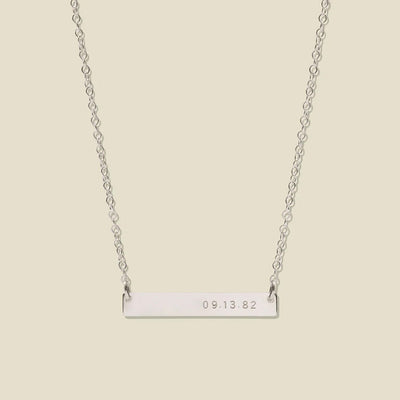 A silver bar necklace with a delicate chain, featuring the date '09.13.82' engraved on the right side of the bar, set against a solid light beige background.
