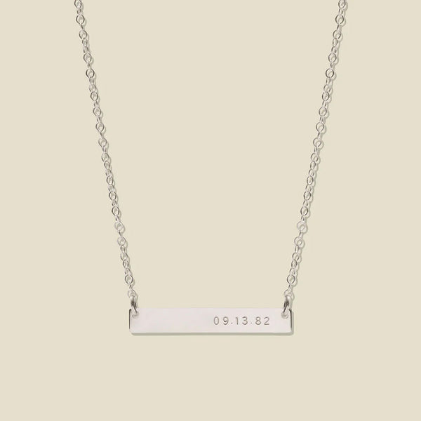 A silver bar necklace with a delicate chain, featuring the date '09.13.82' engraved on the right side of the bar, set against a solid light beige background.