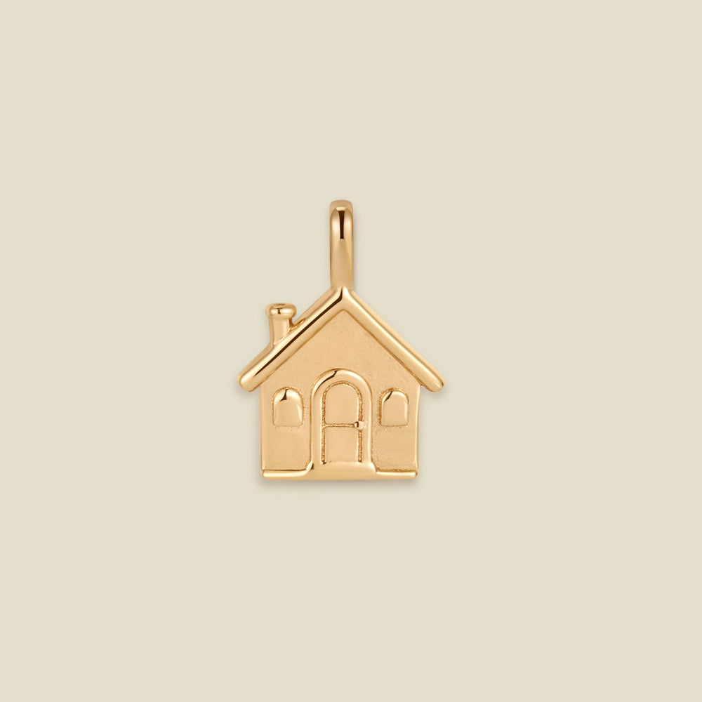 A small gold house-shaped charm pendant with a loop at the top, shown centered on a plain warm beige background.