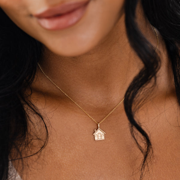 A close-up of a person's neck wearing a delicate gold chain necklace with a gold house charm.