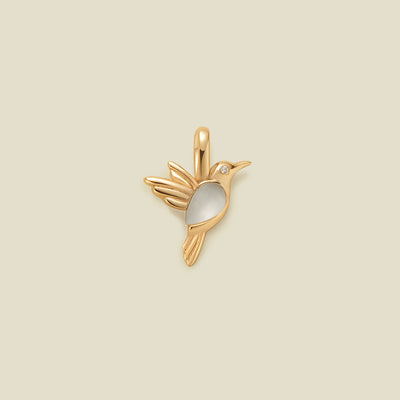 A gold bird charm with a smooth white stone belly and a small clear gemstone eye, centered on a solid beige background.