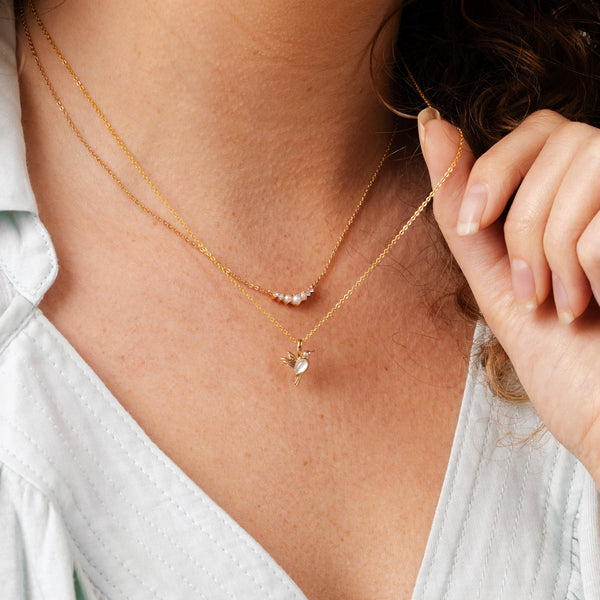 A close-up of a person wearing a delicate gold chain necklace with a crescent shape of pearls and clear gemstones, layered with a delicate gold pendant chain with a gold hummingbird charm.