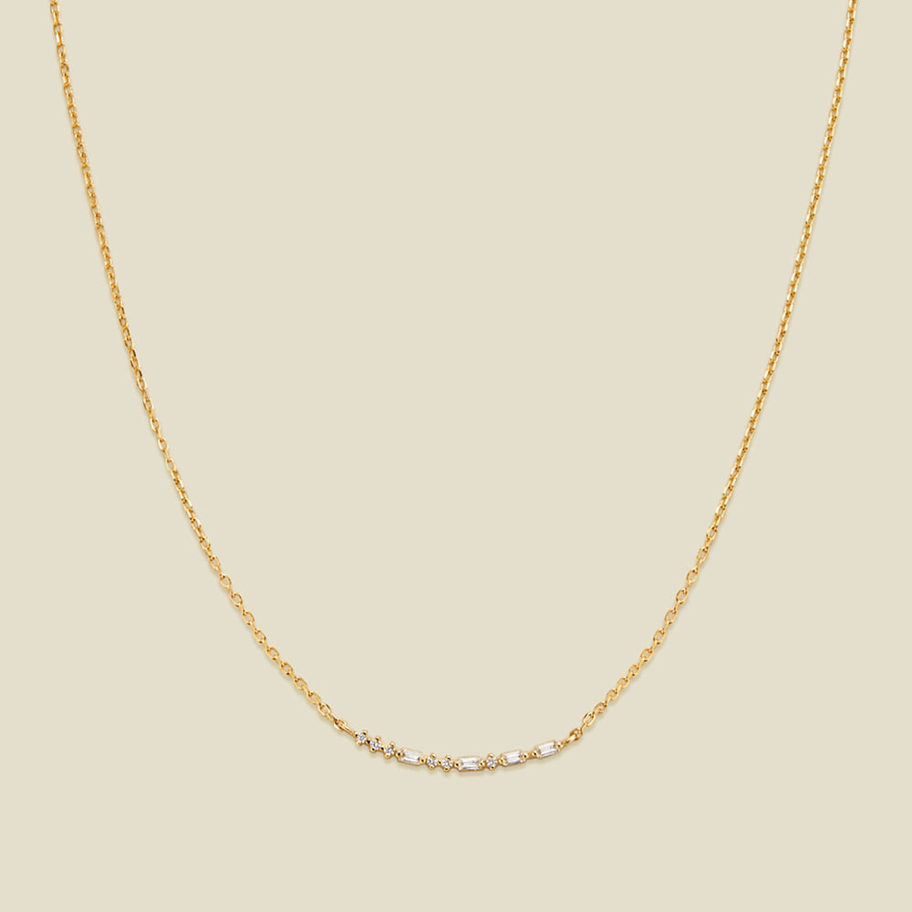 A delicate gold chain necklace featuring a short central crescent pattern with small clear baguette and round cut stones, displayed against a solid light beige background.