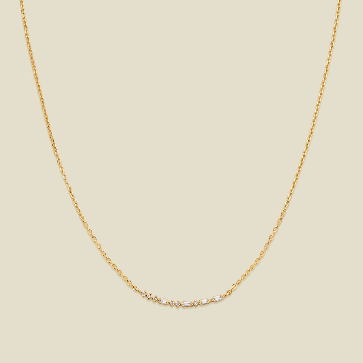 A delicate gold chain necklace featuring a short central crescent pattern with small clear baguette and round cut stones, displayed against a solid light beige background.